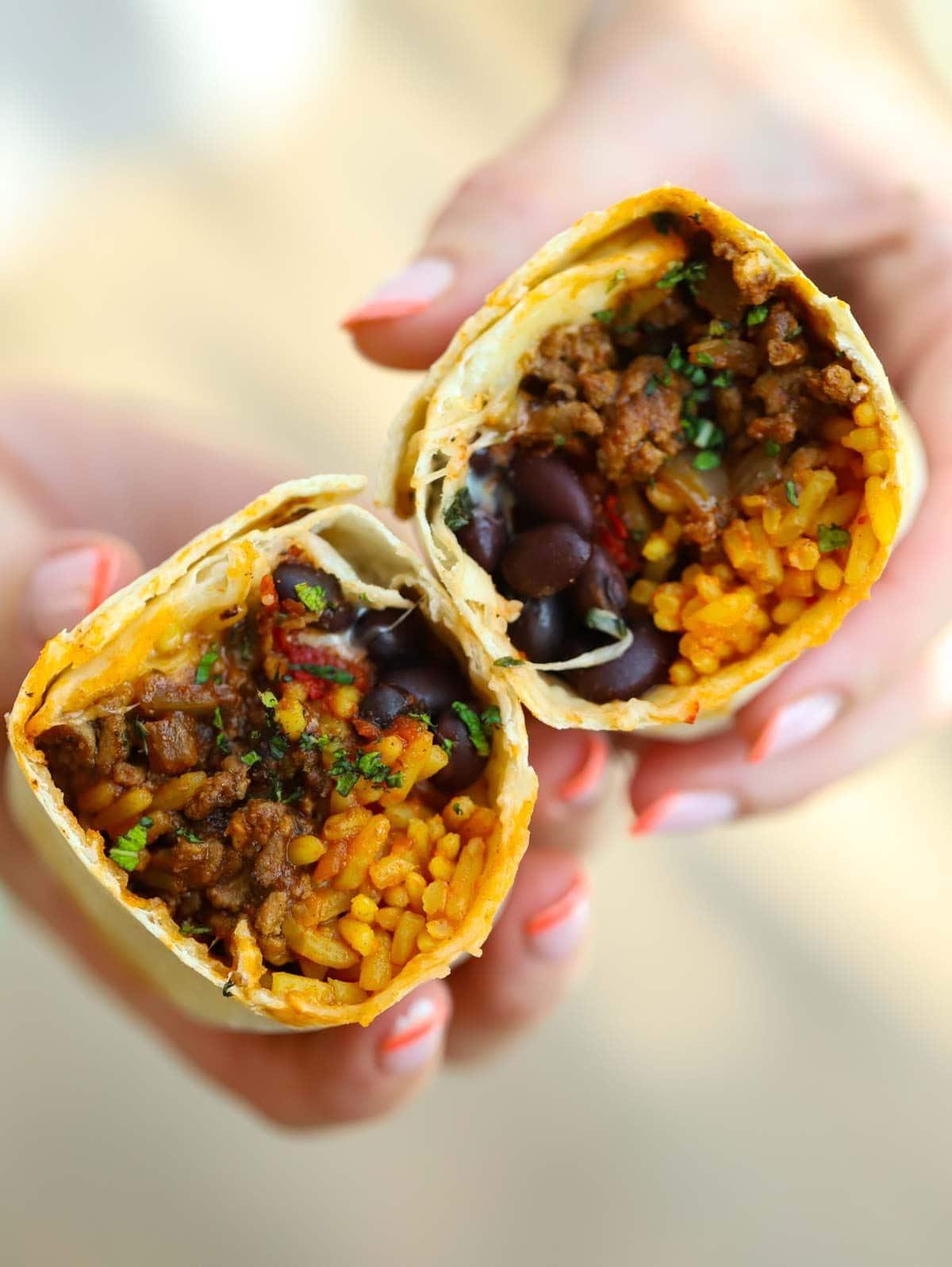 **I Burritos di Manzo Più Semplici di Sempre {Pronto in 30 Minuti}** 2 Due mani che tengono due metà separate di un Burrito di Manzo, ripieno di carne macinata, riso, fagioli e formaggio.