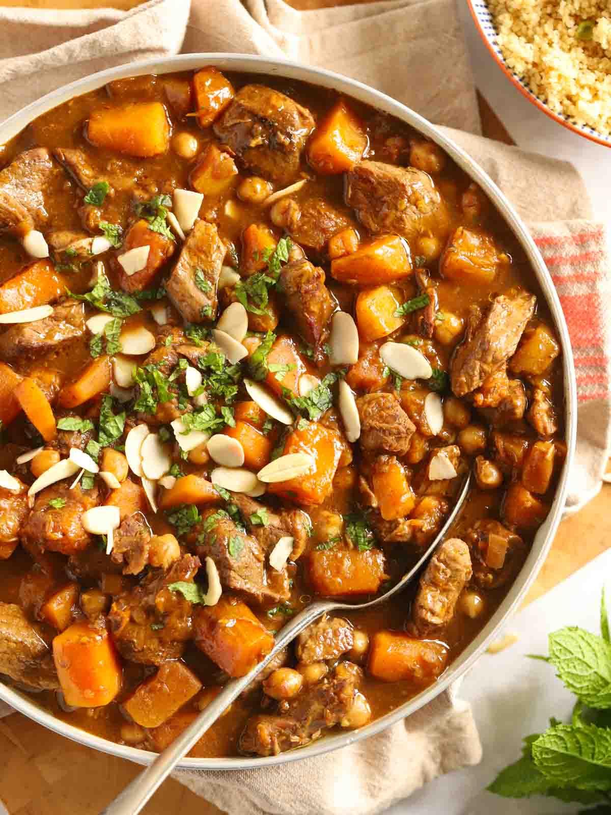 Tagine di Agnello {Facile Stufato alla Marocchina} 2 Una ciotola di tagine di agnello con pezzi di agnello e zucca delica, su un tavolo con un canovaccio, cuscus e un cucchiaio d'argento pronto per essere servito.