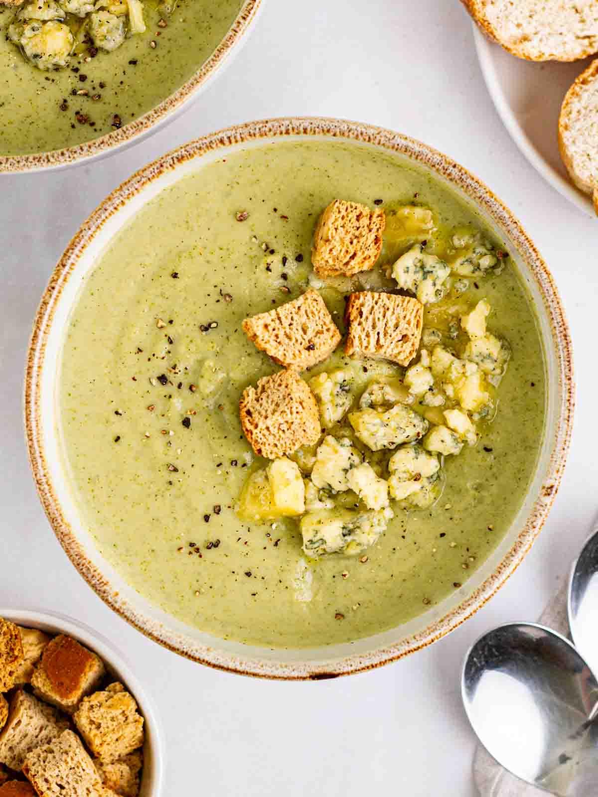 Zuppa di Broccoli e Stilton 2 Una ciotola di Zuppa di Broccoli e Stilton guarnita con formaggio e crostini su un tavolo con zuppa, pane e cucchiai.