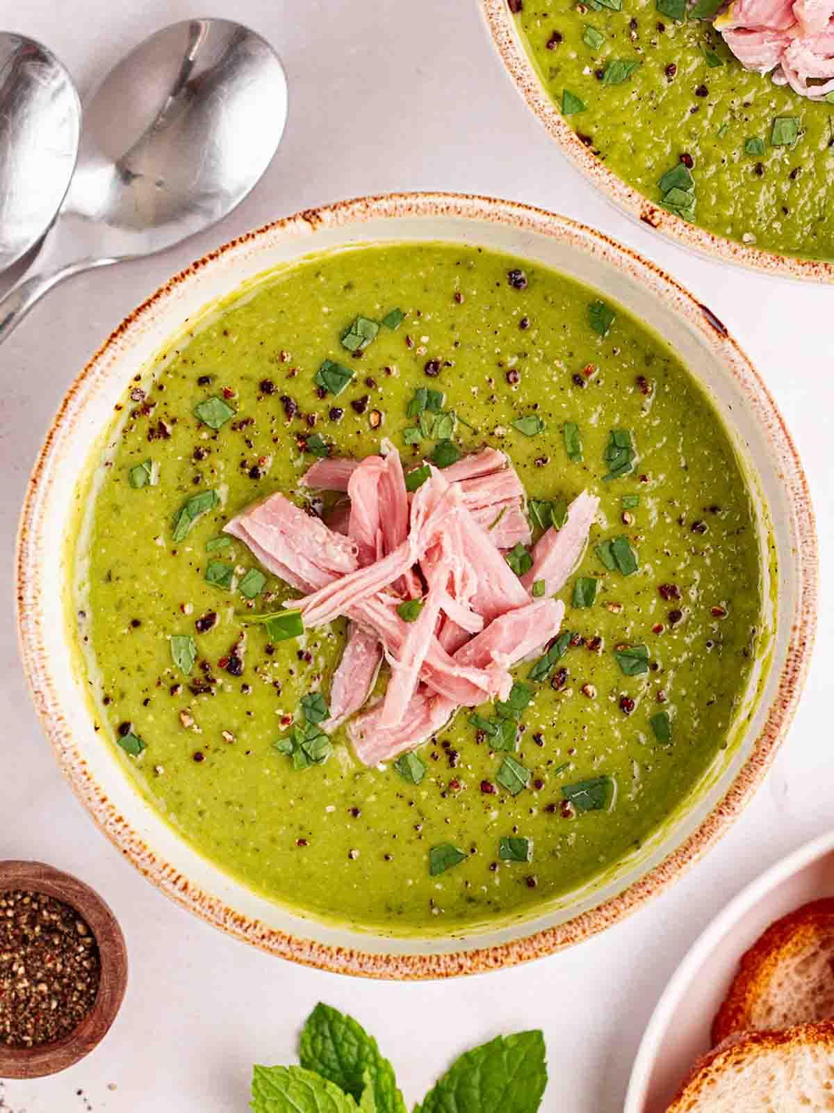 Un piano bianco con ciotole di Zuppa di Piselli e Prosciutto cotta, con cucchiai, pronta da mangiare.