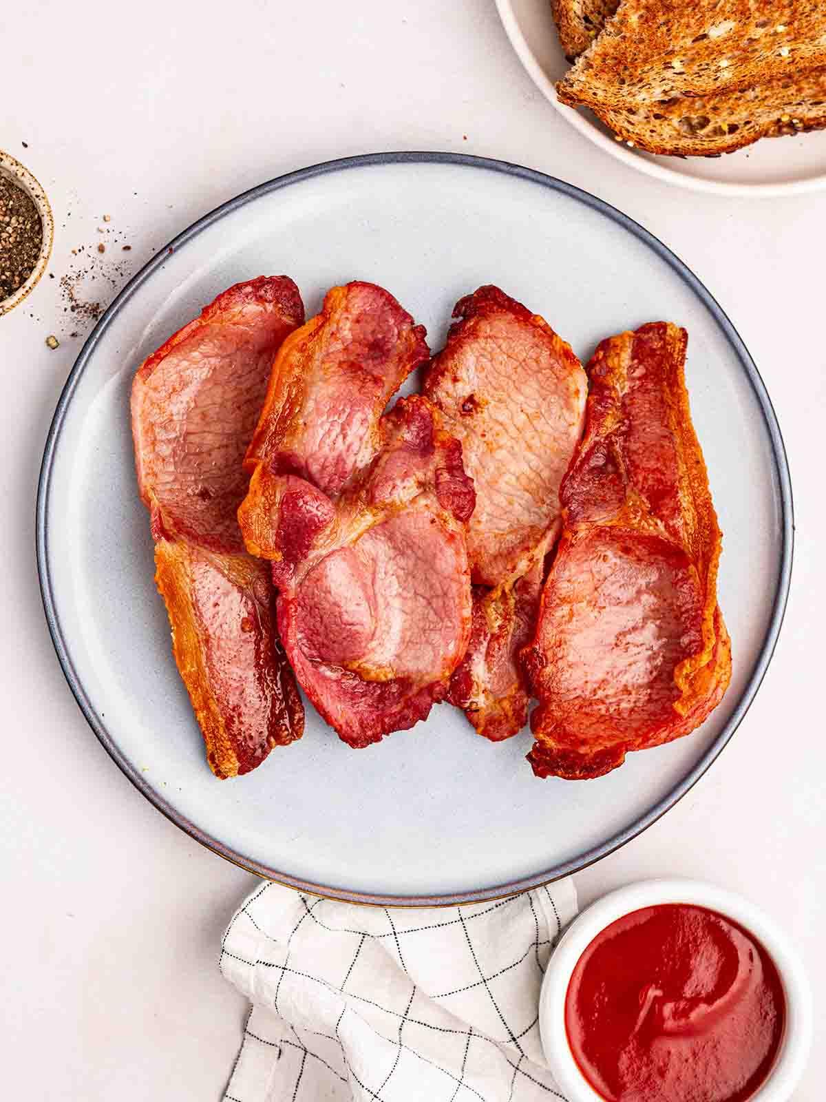 Bacon Perfetto nella Friggitrice ad Aria 2 Un piatto con pancetta cotta nella friggitrice ad aria, su un tavolo con pane tostato a lato, ketchup in una ciotola e pepe nero.