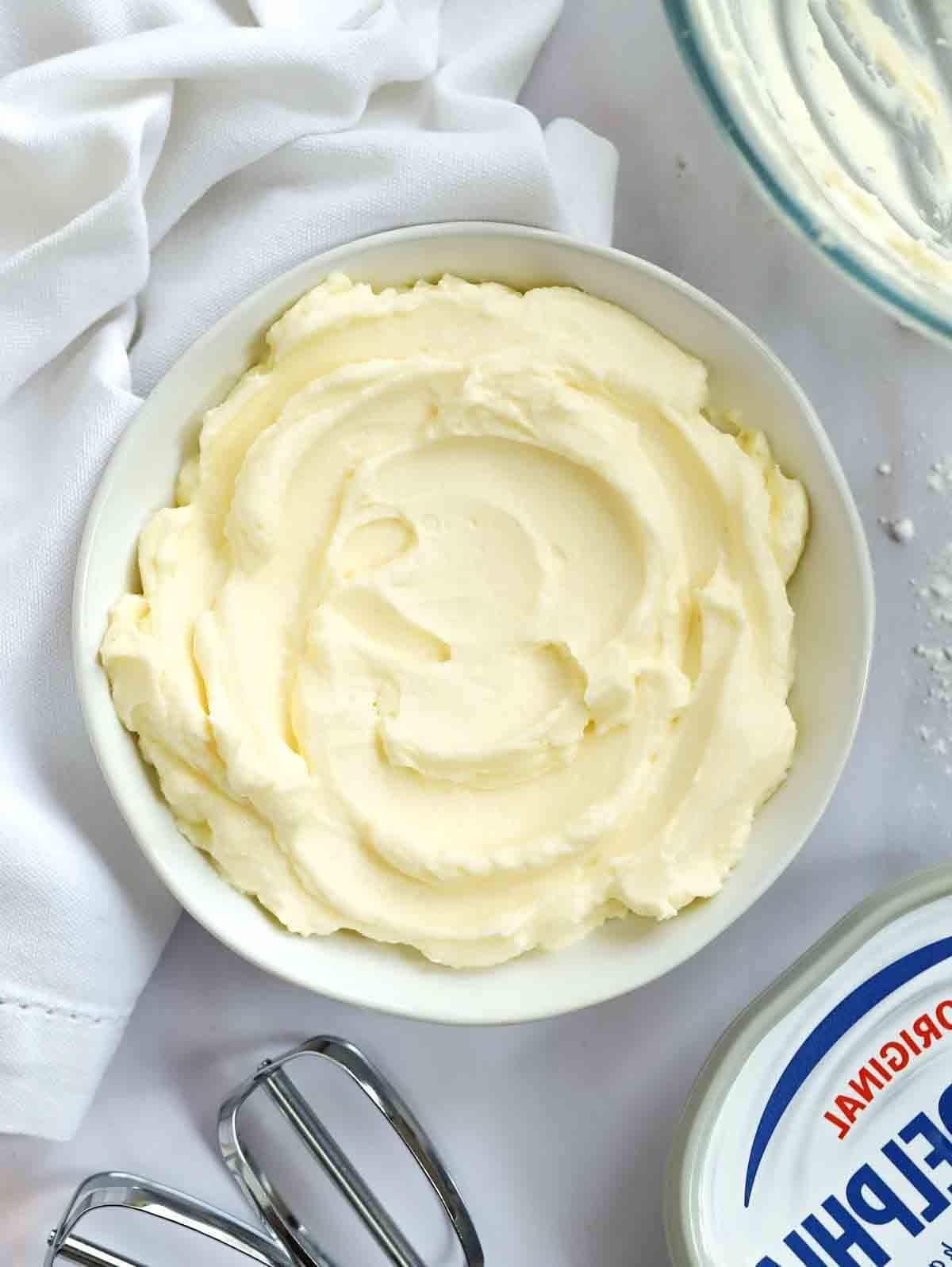 Glassa al Formaggio Cremoso {per Torte e Dolci al Forno} 2 Un recipiente su una tovaglia bianca, pieno di glassa al formaggio cremoso, circondato da una ciotola e una confezione di formaggio spalmabile e una frusta.
