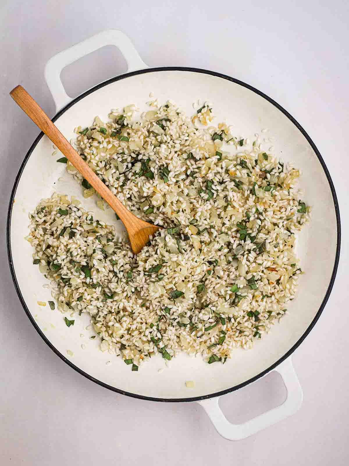 **Risotto di Zucca {Al Forno o Tradizionale}** 11 Una padella con riso per risotto, salvia e cipolla in cottura, con un cucchiaio di legno che mescola.