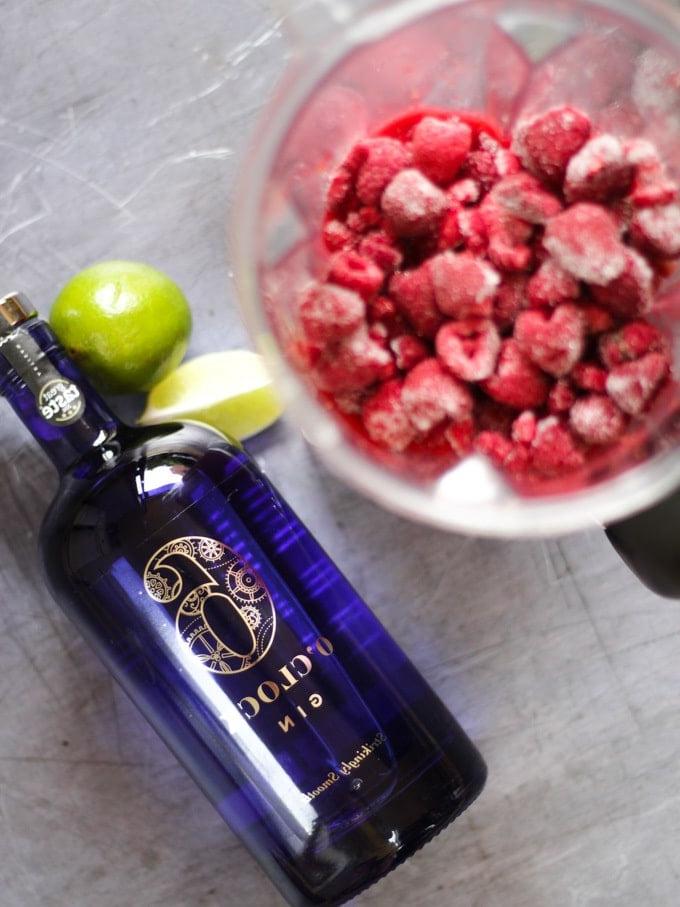 Ripresa dall'alto di lamponi congelati in un frullatore, lime e una bottiglia di gin Waitrose 6 o'clock blu per la ricetta del Cocktail Ghiacciato al Gin e Lamponi.