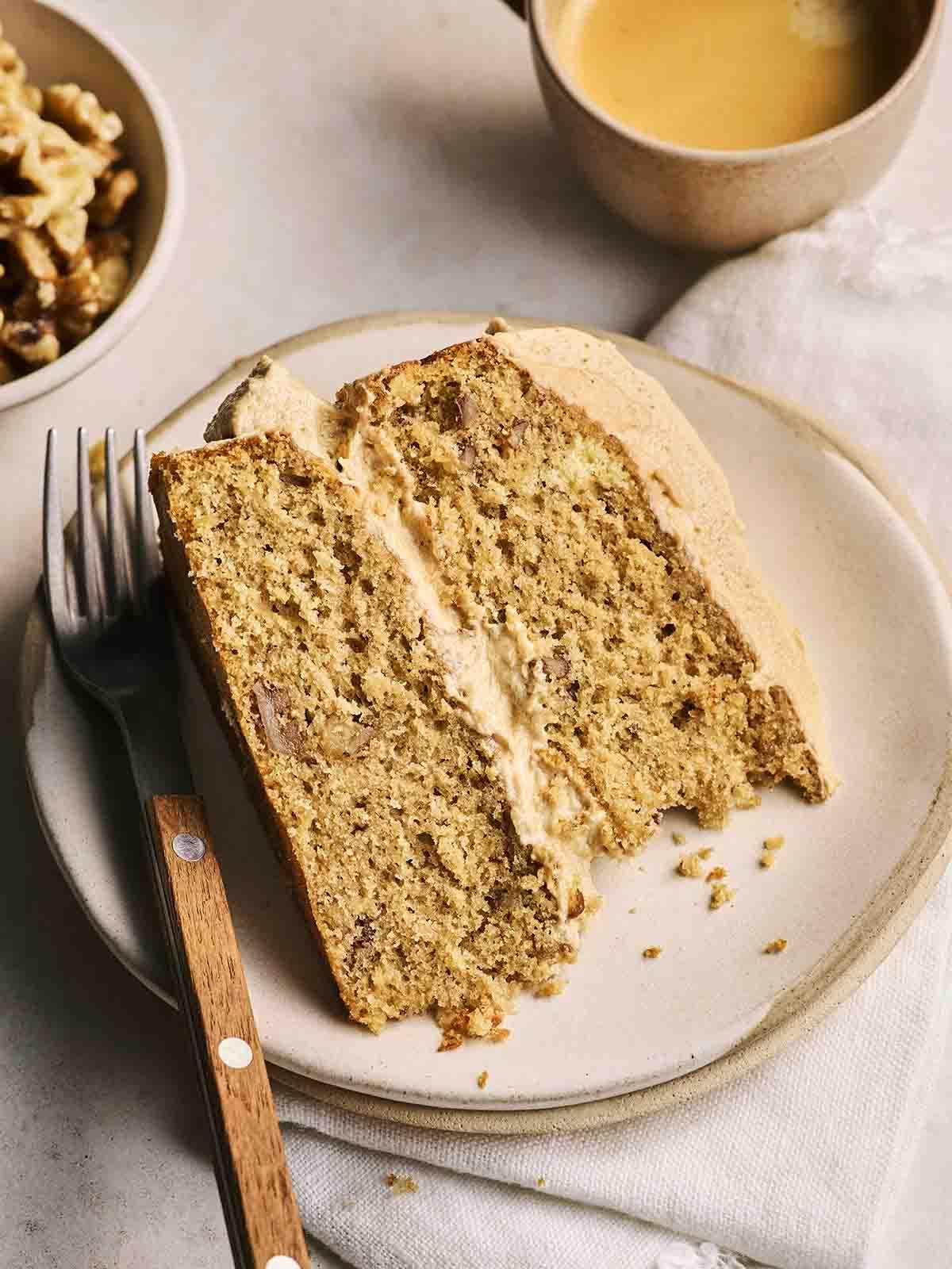 Una fetta di Torta al Caffè e Noci su un piatto bianco, con una forchetta a lato e una tazza di caffè in porcellana sullo sfondo, pronta per essere gustata.