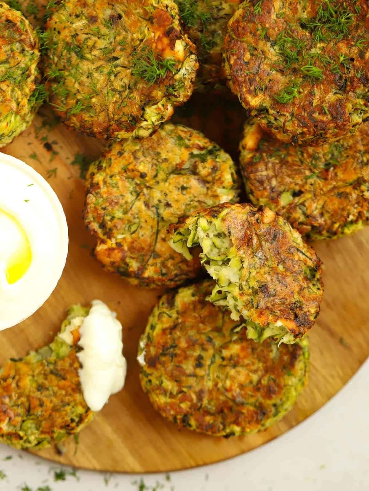 Frittelle di zucchine su un tagliere, con una a cui manca un morso e un altro pezzo intinto in una salsa cremosa.