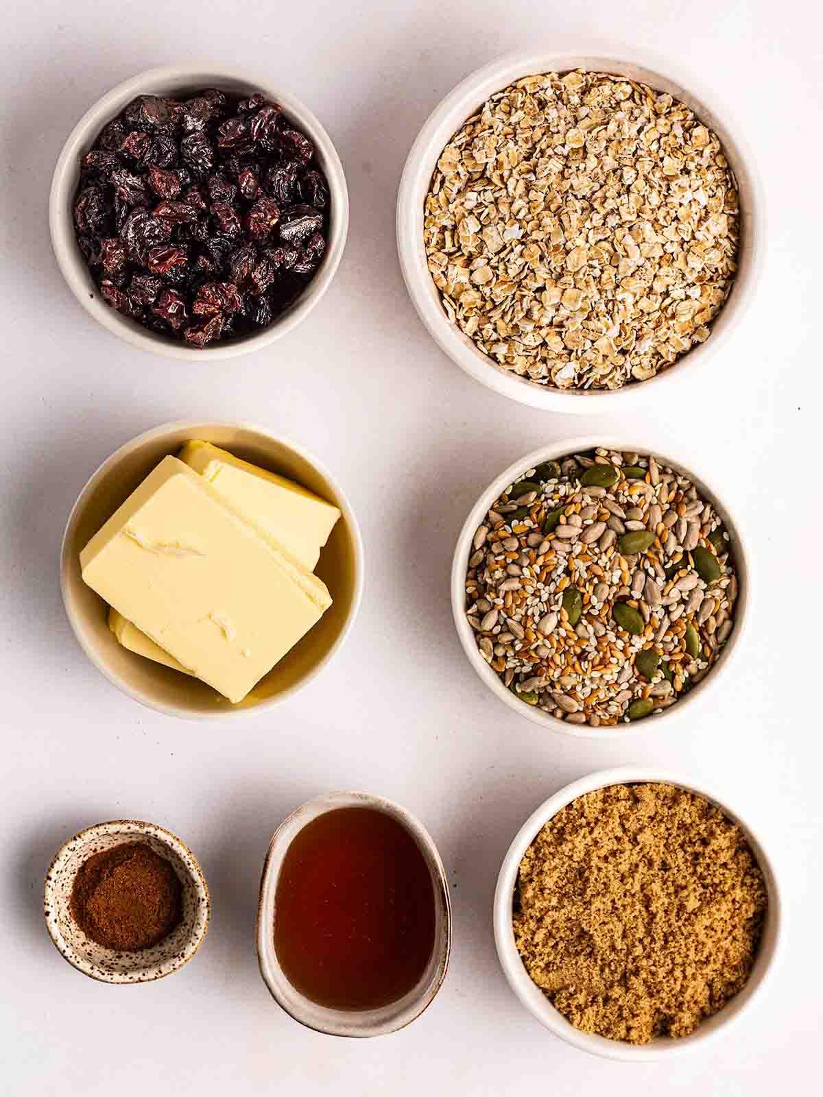 Barrette di Granola {Ricetta Facile!} 3 Gli ingredienti per le barrette di granola fatte in casa disposti in piccole ciotole su un bancone bianco.