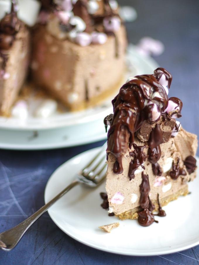 Cheesecake Rocky Road Senza Cottura 3 Fetta di cheesecake Rocky Road senza cottura, cheesecake al cioccolato guarnita con marshmallow e cioccolatini e ricoperta di cioccolato, su un piatto bianco con una forchetta.
