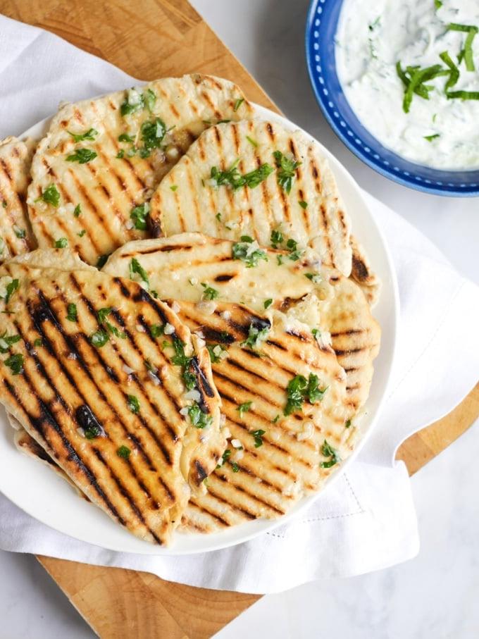 Foto di un piatto di focacce con segni di griglia, su un piatto bianco con tovagliolo bianco e tagliere di legno, sfondo in marmo e salsa tzatziki in una ciotola.
