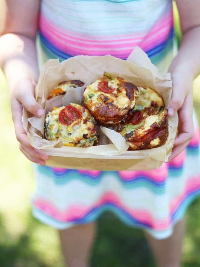 **Ricetta Mini Quiche Senza Crosta** 3 Mini Quiche Senza Crosta con Pomodorini in un cestino per il pranzo foderato con carta da forno, tenuto nelle mani di una bambina, pronta per gustare il suo pasto sano e delizioso.