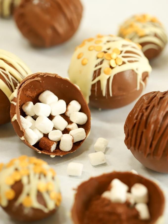 Bombe di Cioccolata Calda {Ricetta Facile} 4 Bombe di cioccolata calda al cacao su una superficie, con una aperta che rivela i marshmallow all'interno.