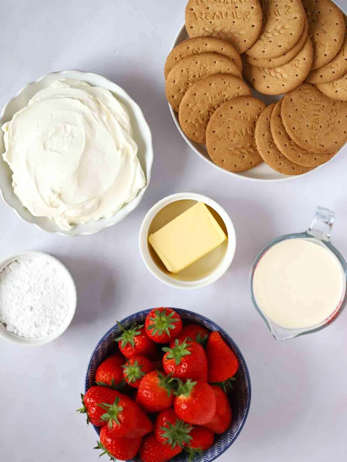Gli ingredienti per una cheesecake alle fragole disposti in ciotole su un bancone bianco.
