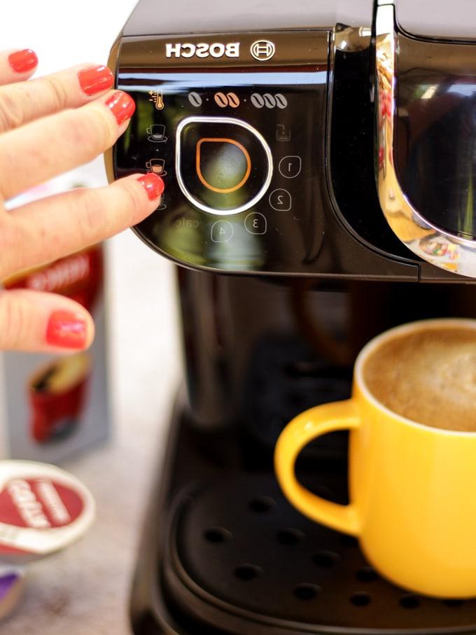 Macchina da caffè Tassimo con dito con unghia rossa che preme un pulsante con tazza gialla piena di caffè e capsule di caffè sullo sfondo per preparare la ricetta del caffè freddo con caramello salato.
