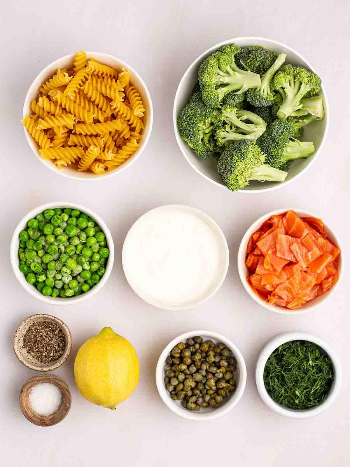 Gli ingredienti per la pasta al salmone affumicato in ciotole su un piano di lavoro bianco.