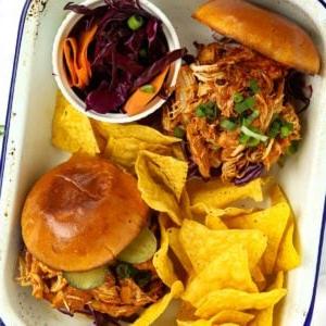Un piatto con due panini ripieni di pollo sfilacciato in slow cooker, pronti da mangiare, con tortilla chips a lato e coleslaw.
