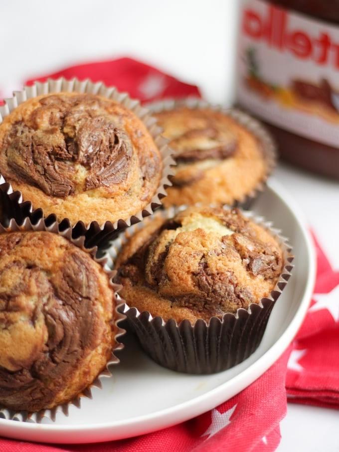 Una pila di muffin alla Nutella in pirottini marroni su un piatto bianco su un panno rosso con stelle bianche e un vasetto di Nutella sullo sfondo.