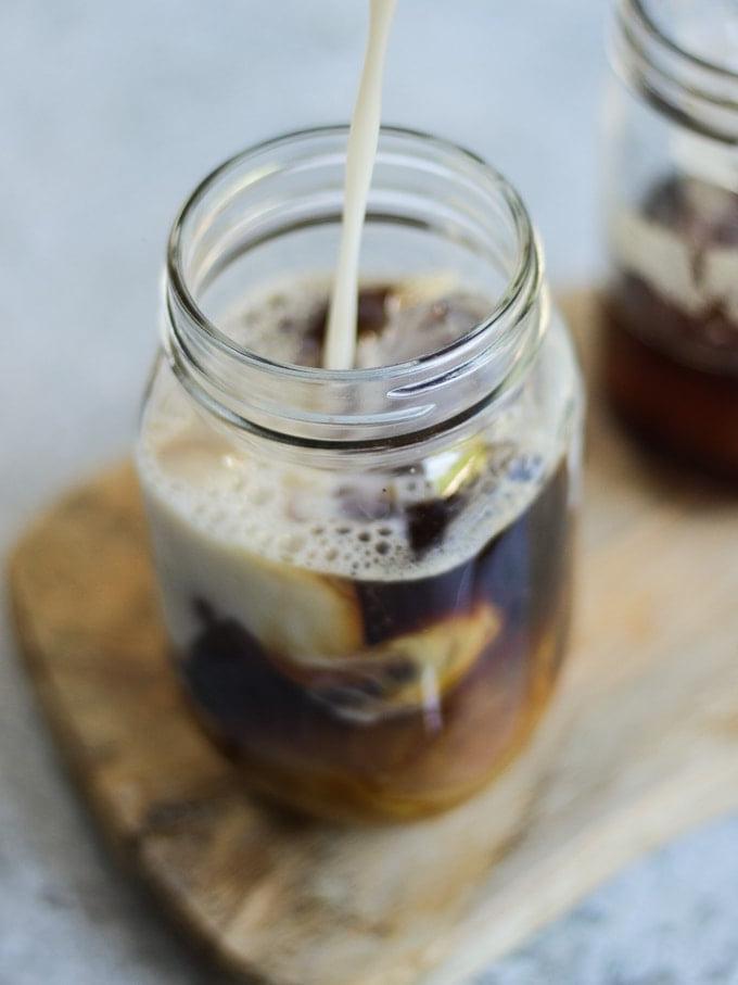 Latte che viene versato nel caffè freddo in una tazza di vetro su un tagliere di legno.