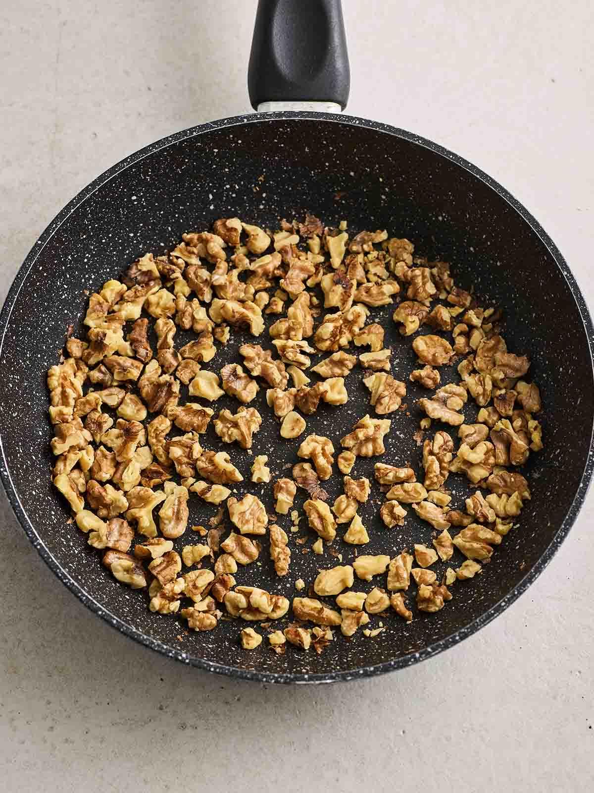 Una padella nera con noci tritate che stanno tostando, per il passo 1 della ricetta dell'Insalata Waldorf.