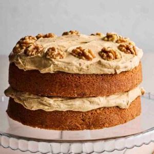Una vista laterale di una classica Torta al Caffè e Noci, con due strati di pan di Spagna e abbondante crema al burro al caffè e noci sulla superficie, posta su un'elegante alzata di vetro.