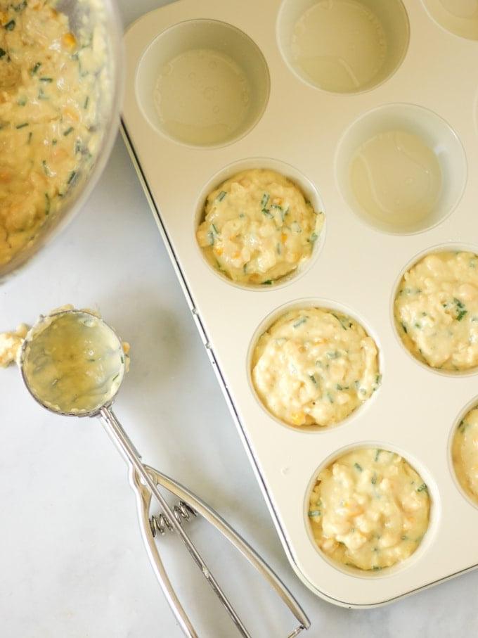 Muffin Salati {Formaggio e Mais} 5 Una ciotola di impasto per muffin salati e un porzionatore da gelato, con una teglia da muffin riempita a metà con l'impasto.