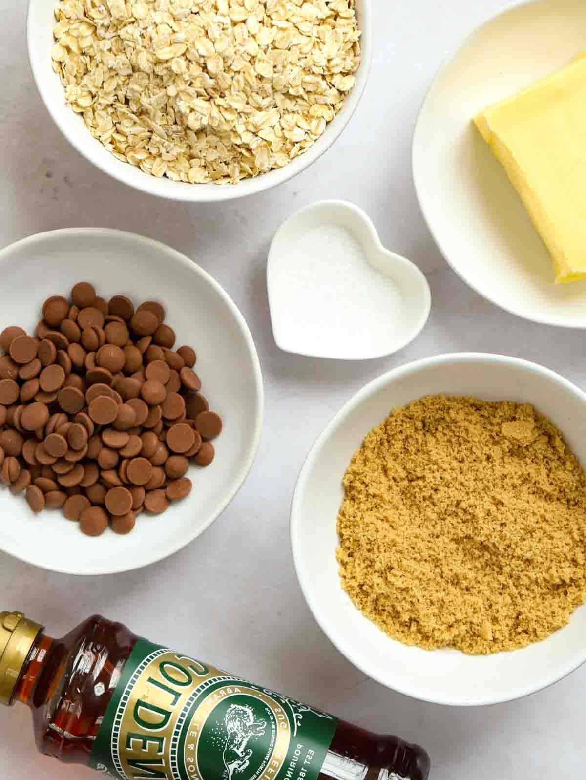 Pancakes Facili {Ricetta Infalibile!} 5 Gli ingredienti grezzi per una ricetta di flapjack disposti su un bancone, inclusa una bottiglia di Golden Syrup e ciotole piene di avena, burro, zucchero, sale e gocce di cioccolato.