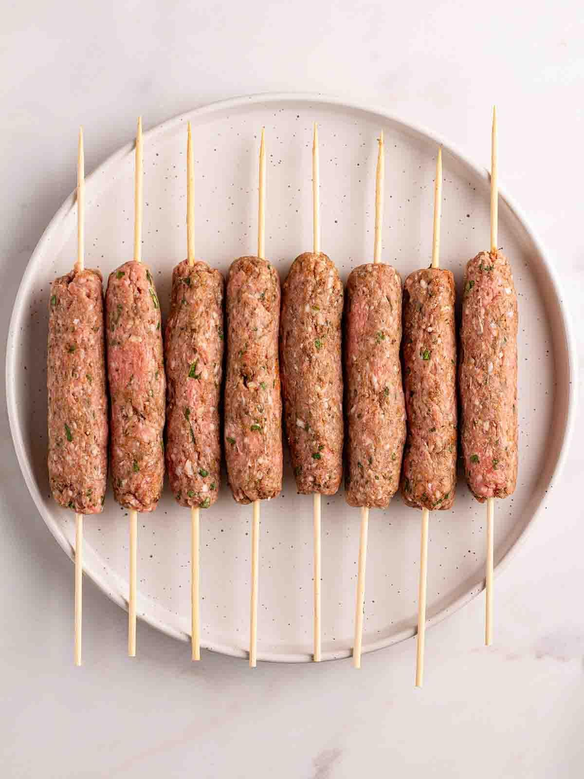 Facili Kofta di Agnello {con Tzatziki} 5 Spiedini di Kofta di agnello crudi con gli spiedini al centro, su un piatto, pronti per essere cotti.