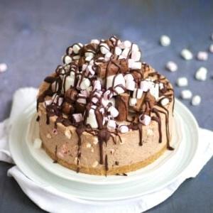 Cheesecake Rocky Road Senza Cottura 6 Una Cheesecake Rocky Road su un piatto bianco, riccamente guarnita con marshmallow e barrette di cioccolato e ricoperta di cioccolato fuso.