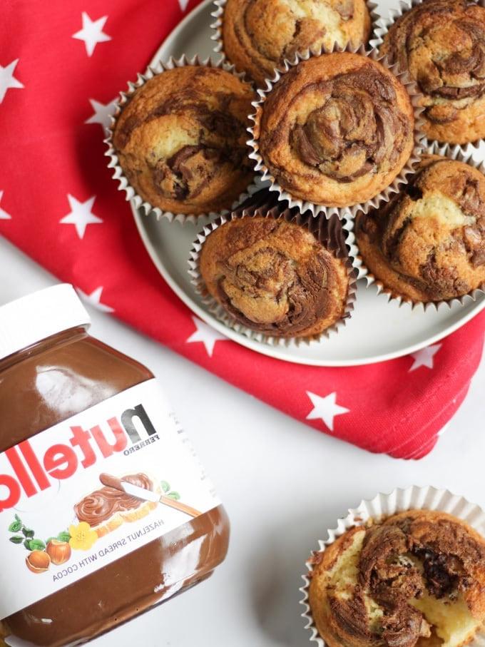 Pila di muffin alla Nutella su un piatto con un panno rosso a stelle bianche sotto, un muffin aperto e un vasetto di Nutella.
