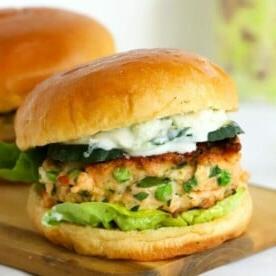 Piano Pasti 7 6 Hamburger di salmone freschi in un panino, serviti su un tagliere di legno, accompagnati da salsa tzatziki.