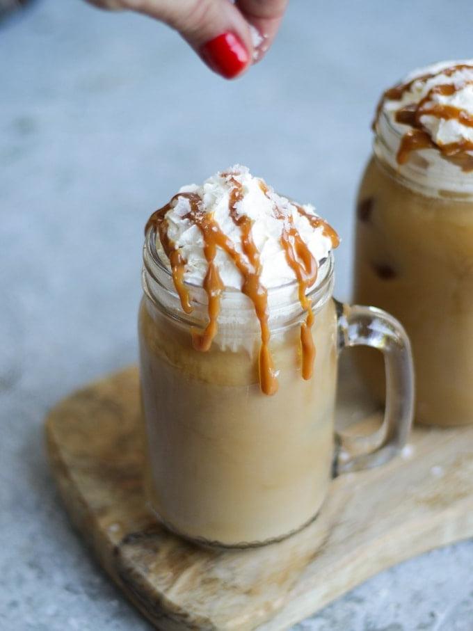 Due tazze di vetro con manico di caffè freddo con caramello salato e capsule Tassimo sullo sfondo grigio con dita che spargono sale sulla parte superiore con unghie rosse.