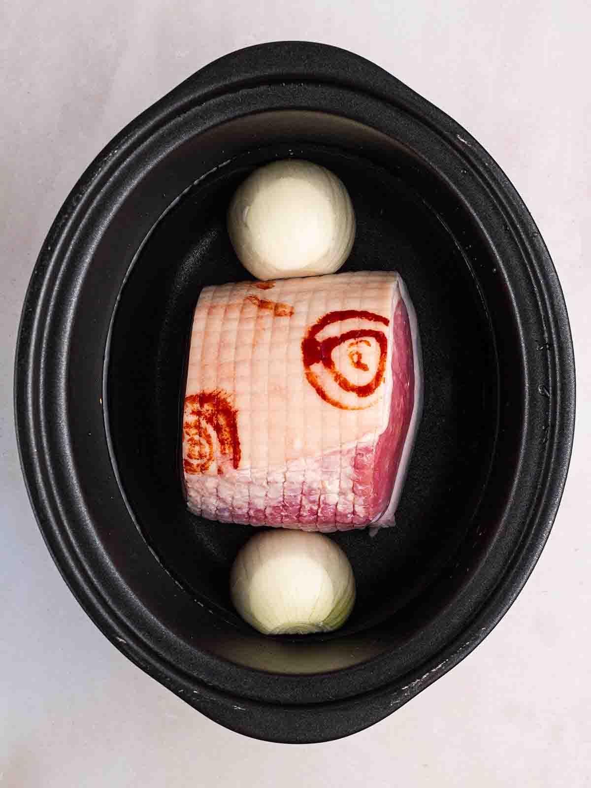 Una slow cooker riempita con un taglio di gammon, cipolla e acqua per il passo 1 della ricetta per la Zuppa di Piselli e Prosciutto.