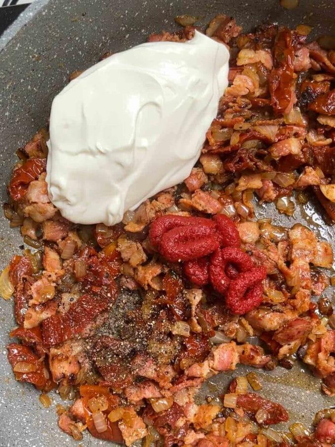 Una cucchiaiata di formaggio cremoso aggiunta a una padella di pomodori secchi fritti, cipolla e pancetta per la ricetta della Pasta con Pancetta. Passaggio 3.