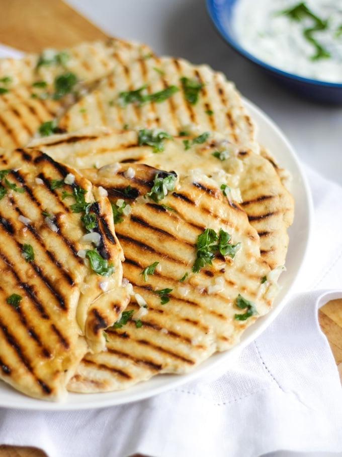Foto laterale di un piatto di focacce facili con segni di griglia, su un piatto bianco con tovagliolo bianco e tagliere di legno, sfondo in marmo e salsa tzatziki in una ciotola.