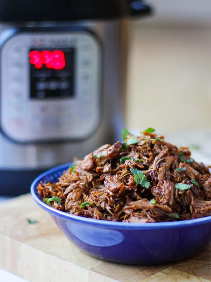 Ciotola blu piena di maiale sfilacciato Instant Pot con Instant Pot sullo sfondo.