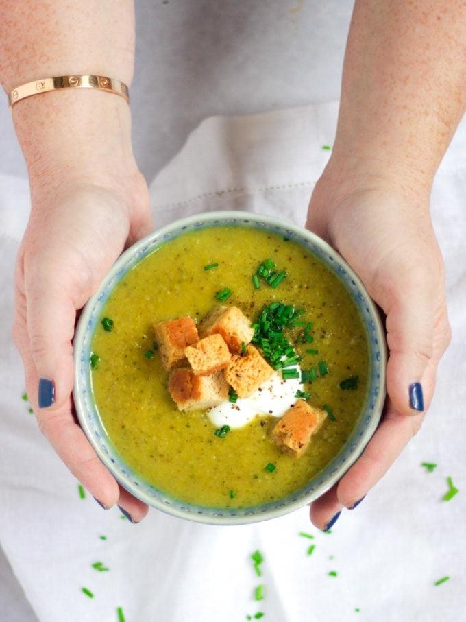 Zuppa Facile di Zucchine al Parmigiano 7 Una ciotola blu di vellutata di zucchine tenuta in mano con panna acida, crostini e erba cipollina su sfondo bianco.