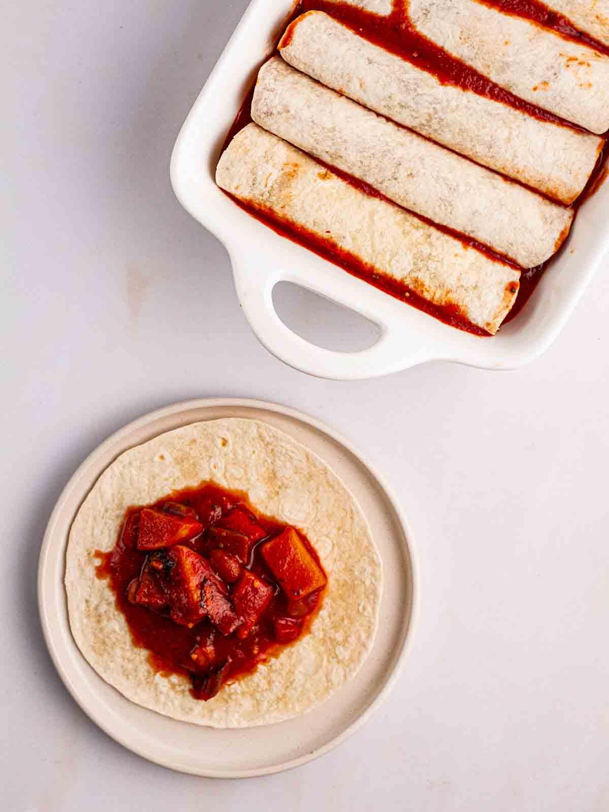 Enchiladas Vegetariane con Zucca Violina 8 Enchiladas arrotolate in una teglia da forno con una tortilla non arrotolata su un piatto per mostrare il ripieno di verdure per il passaggio 4 della ricetta delle Enchiladas Vegetariane.