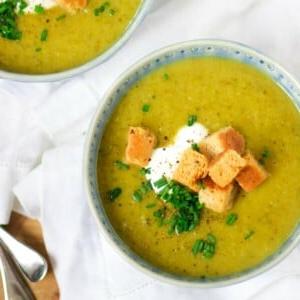 Zuppa Facile di Zucchine al Parmigiano 8 Una ciotola di vellutata di zucchine con guarnizioni di erba cipollina, panna acida e crostini, con un'altra ciotola leggermente inquadrata, entrambe su una tovaglia bianca.