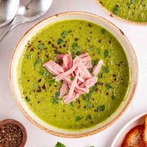 Una pentola di Zuppa di Piselli e Prosciutto verde con prosciutto sfilacciato in cima, pronta da mangiare.