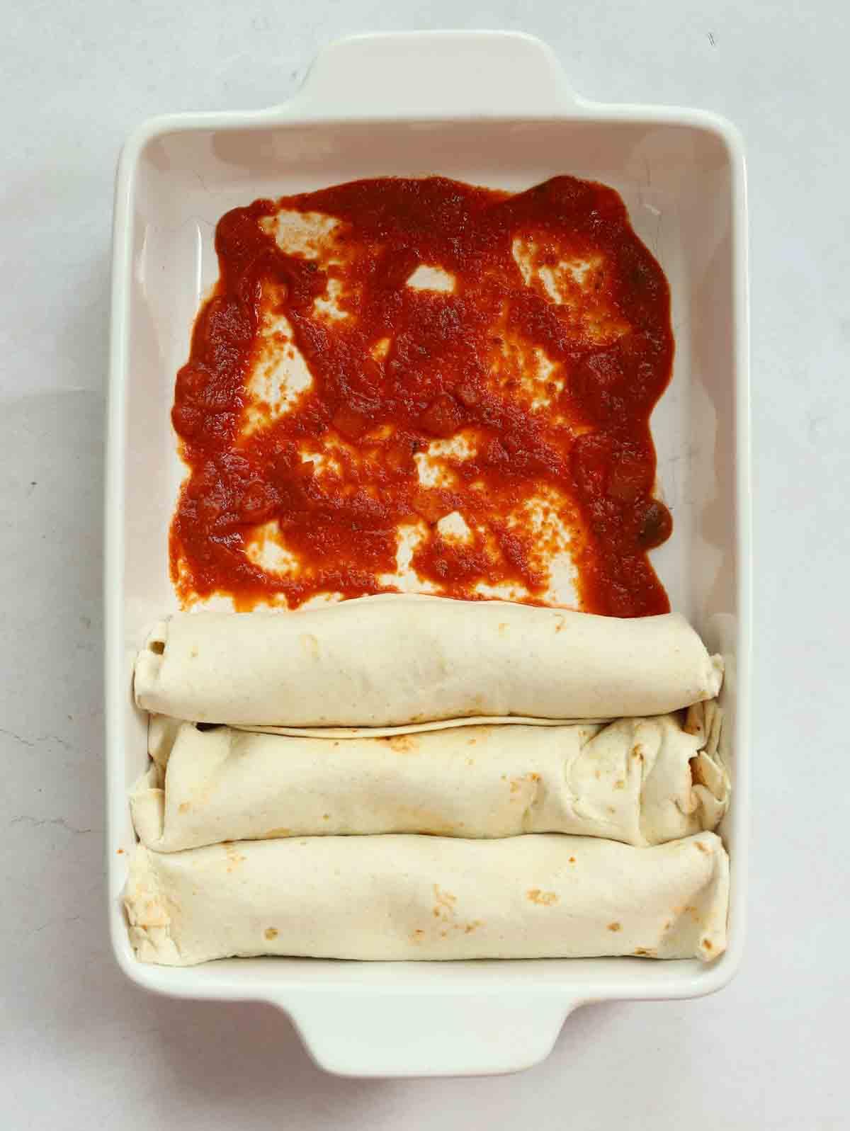 Enchiladas di Pollo 9 Tre enchiladas di pollo arrotolate su un letto di salsa di pomodoro in una teglia adatta al forno.