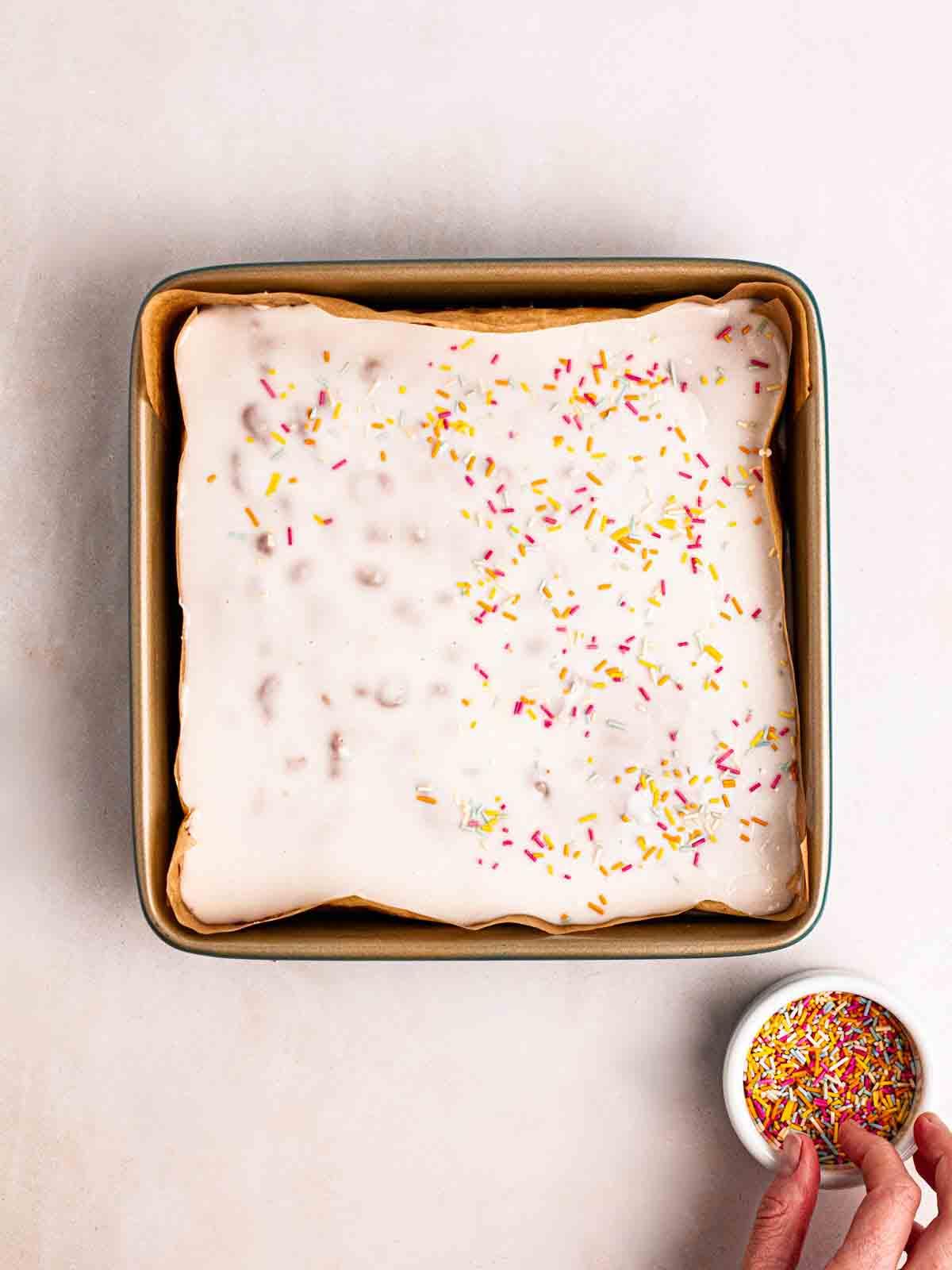 Torta Scolastica {Ricetta Facile!} 10 Una mano che sparge zuccherini su una torta con glassa bianca.