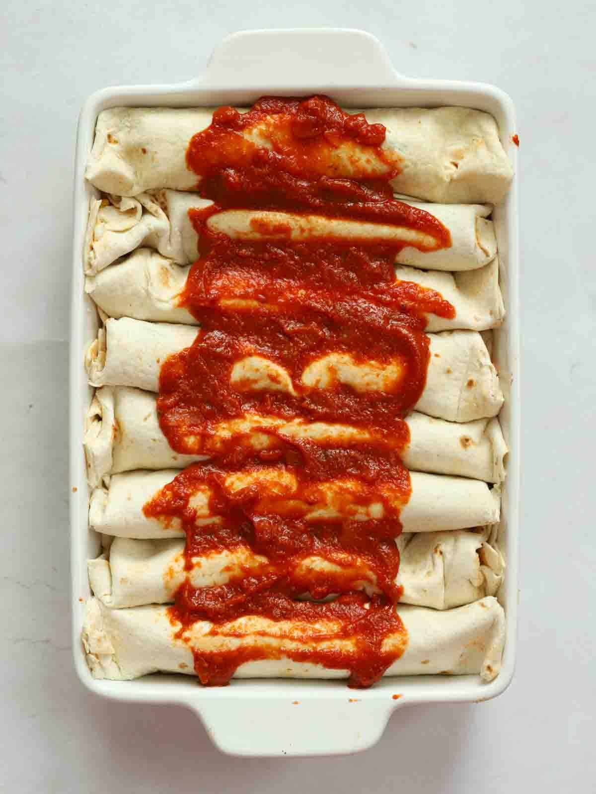 Enchiladas di Pollo 10 Enchiladas di pollo arrotolate in una teglia adatta al forno con salsa di pomodoro sopra, pronte per essere infornate.