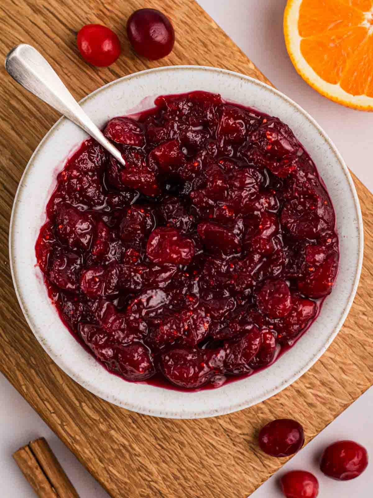 Salsa di Mirtilli Rossi Fatta in Casa 2 Una tavola con una ciotola bianca piena di salsa ai mirtilli rossi, con mirtilli e un'arancia a lato.