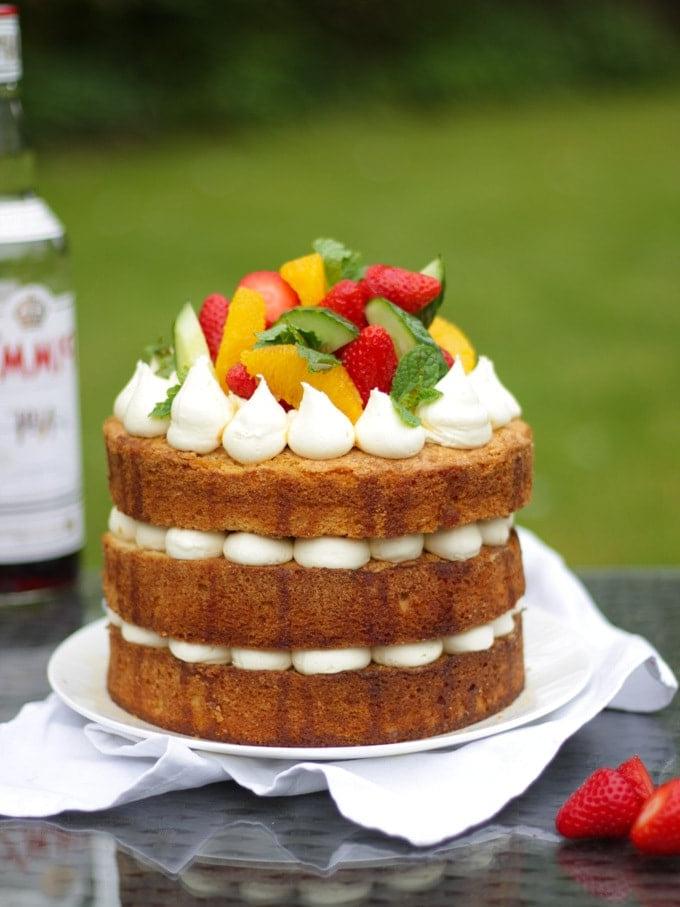 Pimms cake con glassa, cetriolo, fragole, arance e menta su un piatto bianco in giardino, ricetta torta al Pimms.