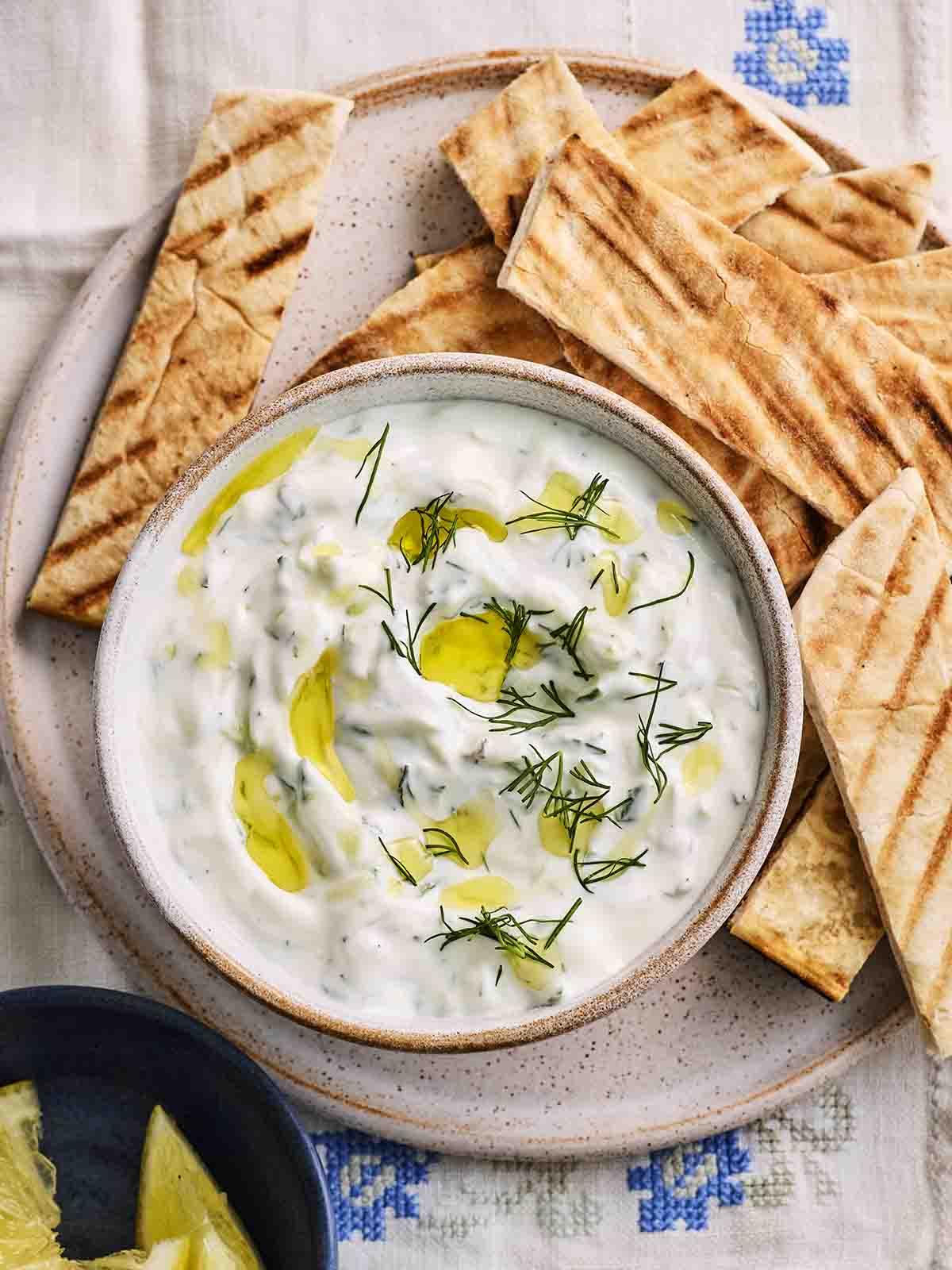Tzatziki {Ricetta Facile!} 2 Una ciotola di Tzatziki greco con olio d'oliva ed erbe fresche in superficie, affiancata da fette di pita.