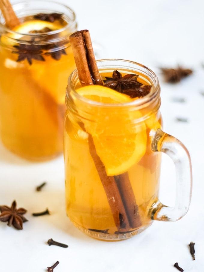 Sidro Caldo Speziato {Ricetta Facile per Slow Cooker} 2 Foto dall'alto di due tazze di vetro di sidro caldo speziato preparato nella pentola a cottura lenta su sfondo bianco, con fette d'arancia, bastoncini di cannella, anice stellato e chiodi di garofano.