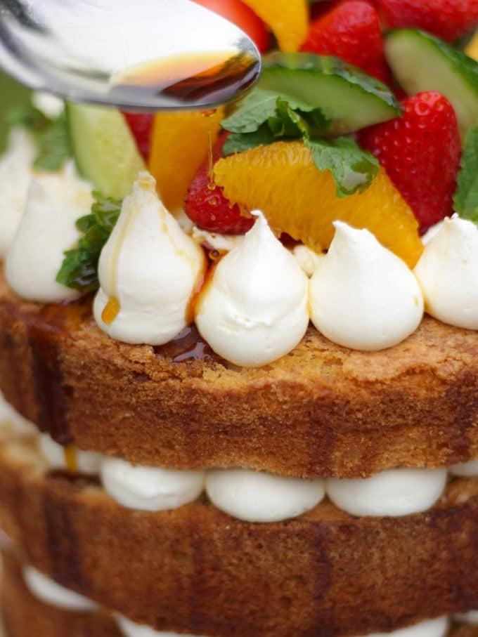 Pimms cake con cucchiaio che irrora sciroppo al Pimms, glassa, cetriolo, fragole, arance e menta su un piatto bianco in giardino, ricetta torta al Pimms.