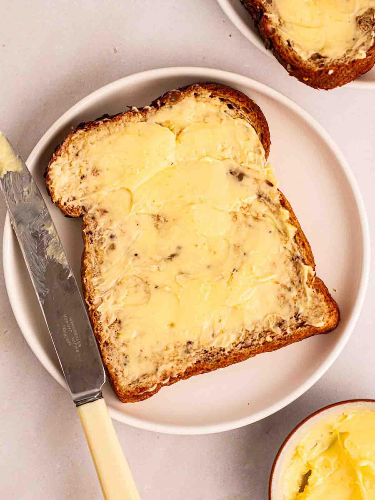 Burro Fatto in Casa {Facile e Delizioso} 3 Burro fatto in casa spalmato densamente su una fetta di pane integrale, su un piatto bianco con un coltello di lato.