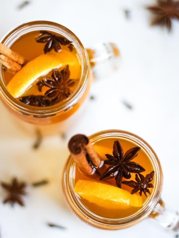 Sidro Caldo Speziato {Ricetta Facile per Slow Cooker} 3 Foto dall'alto di due tazze di vetro di sidro caldo speziato preparato nella pentola a cottura lenta su sfondo bianco, con fette d'arancia, bastoncini di cannella, anice stellato e chiodi di garofano.