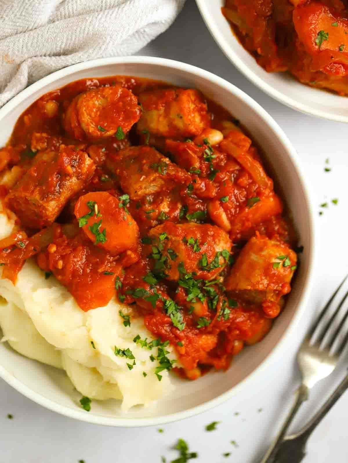 Stufato di Salsicce a Cottura Lenta {Ricetta Facile} 3 A bowl of sausage casserole and mashed potato, laid on a white table with a fork at the side.