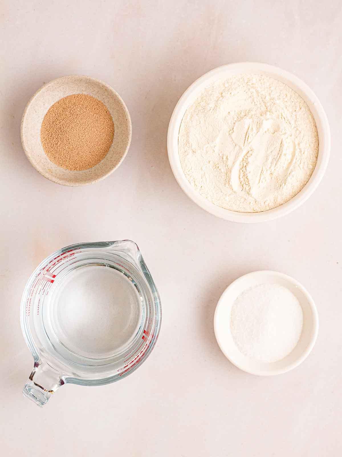 Pane Senza Impasto {La Ricetta Notturna Più Semplice di Sempre} 4 Gli ingredienti per la ricetta del Pane Senza Impasto disposti in ciotole su un bancone bianco.
